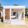 Una piccola e moderna casa prefabbricata con una veranda e sedie, circondata da un prato e alberi.