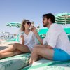 Due persone su una spiaggia, sedute su lettini, mangiano gelato sotto ombrelloni verdi e bianchi.