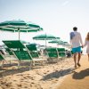 Coppia cammina sulla spiaggia tra lettini e ombrelloni verdi e bianchi.