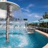 Una fontana a cascata in una piscina, con cielo blu e ombrelloni a righe sullo sfondo.