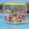 Bambini sorridenti con cappelli colorati giocano sotto una cascata in piscina.